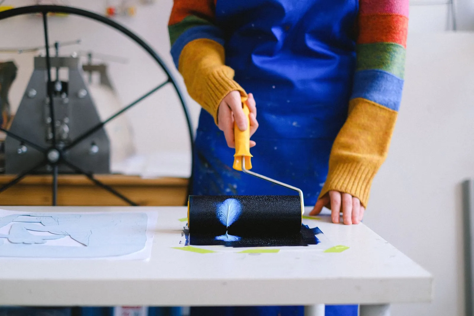 Crop anonymous female artist in casual outfit and dirty apron standing near table while rolling on paper roller covered in paint in light studio
